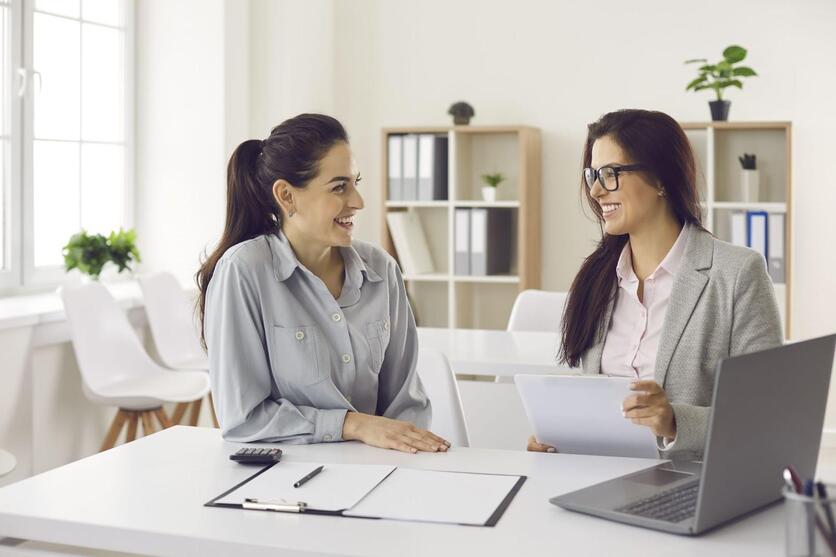 Duas mulheres sentadas no mesmo lado de uma mesa se olhando. Na mesa possui um computador, e uma das mulheres segura uma prancheta.