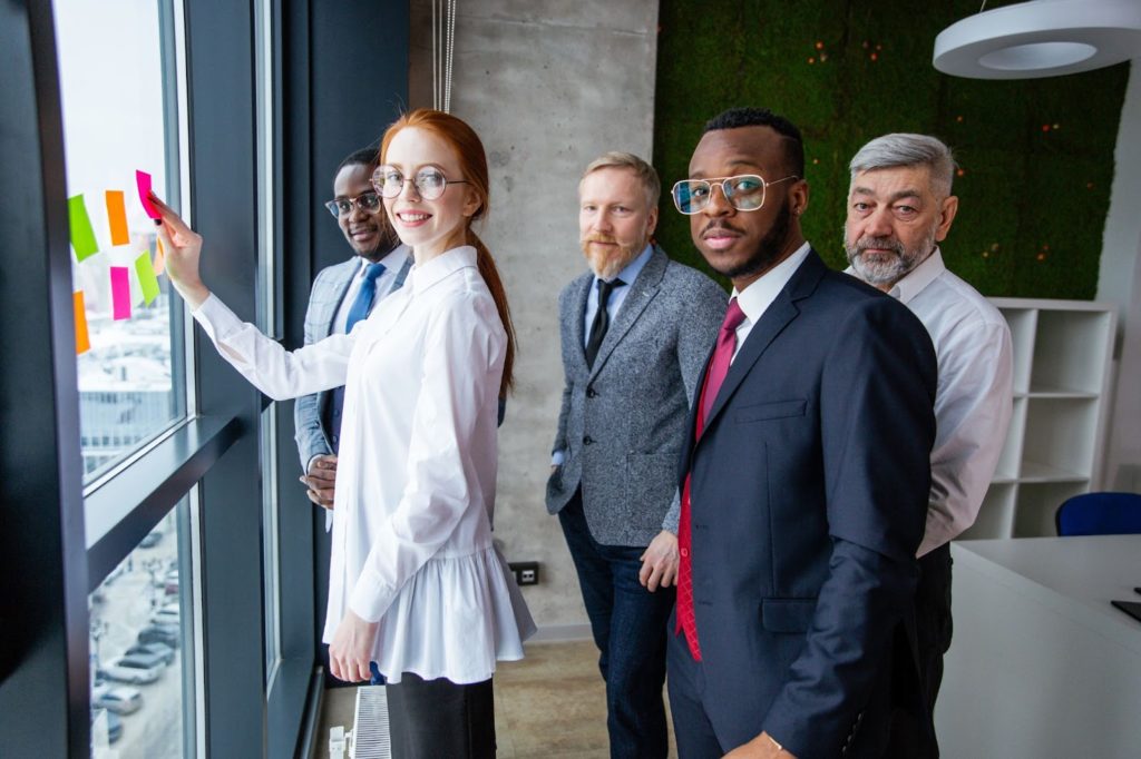 Um grupo de pessoas em frente a uma janela, enquanto olham para a câmera. O ambiente ao redor é corporativo. Uma mulher está colando notas adesivas na janela.