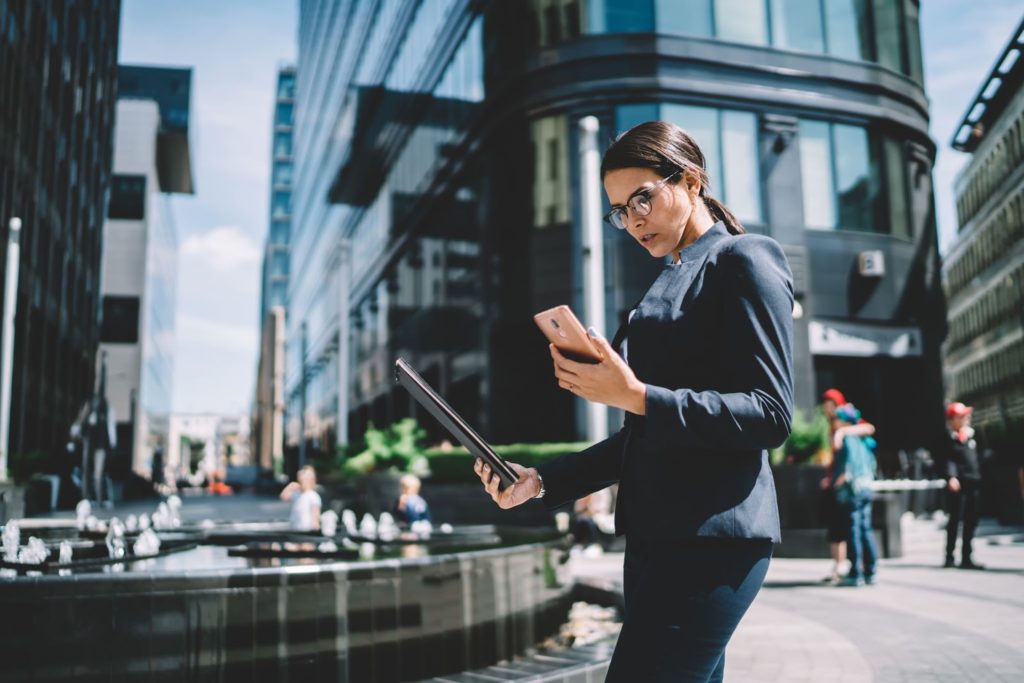 Uma mulher segurando um celular em uma mão e um caderno na outra. Ela está parada na rua, com diversos prédios em volta. 
