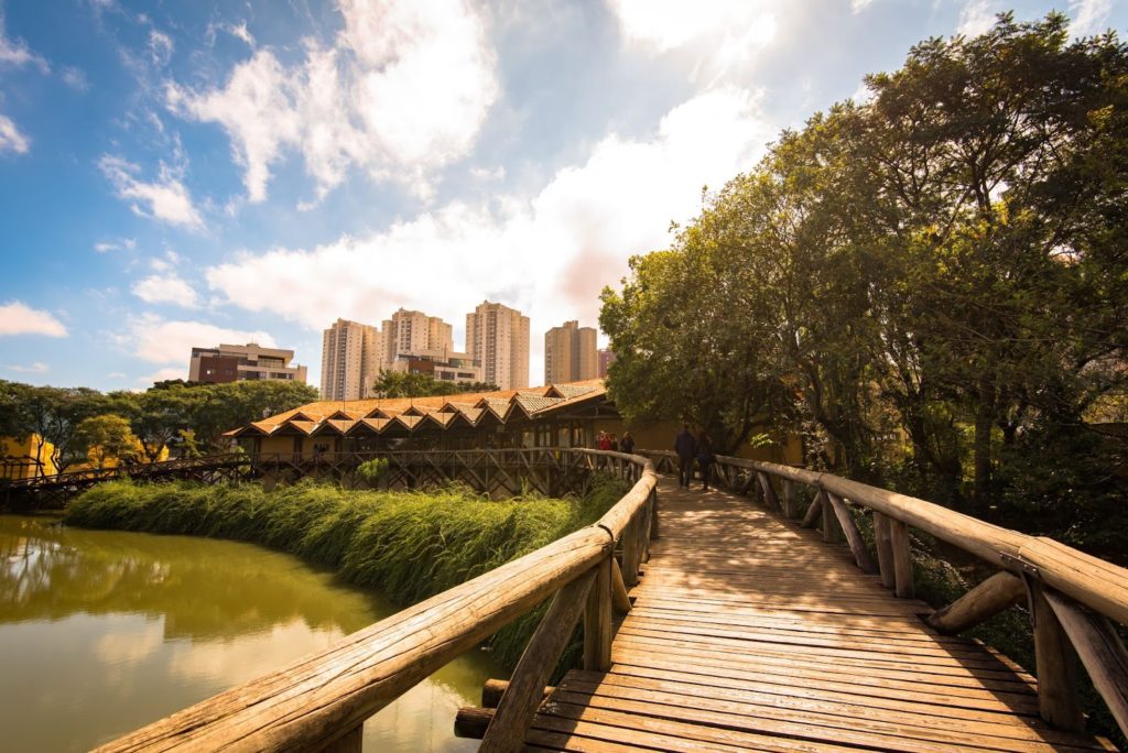 A imagem apresenta uma ponte feita de madeira sobre uma lagoa em um parque de Curitiba, muitas árvores ao lado e também algumas pessoas caminhando. Ao fundo, prédios e o céu ensolarado. 