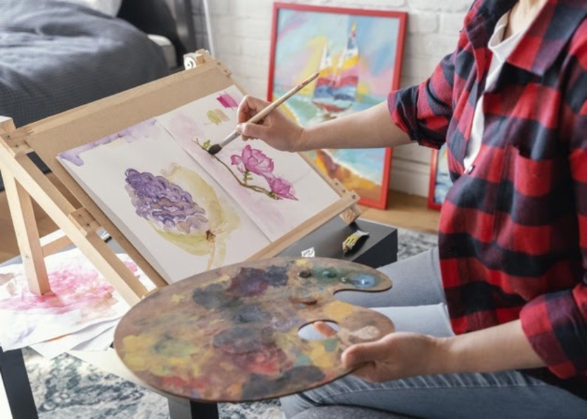 Mulher sentada segurando uma paleta com variadas cores de tinta com a mão esquerda, com a mão direita ela segura um pincel. A sua frente um cavalete, ela está pintando uma rosa. Atrás dela é possível quadros apoiados na parde.