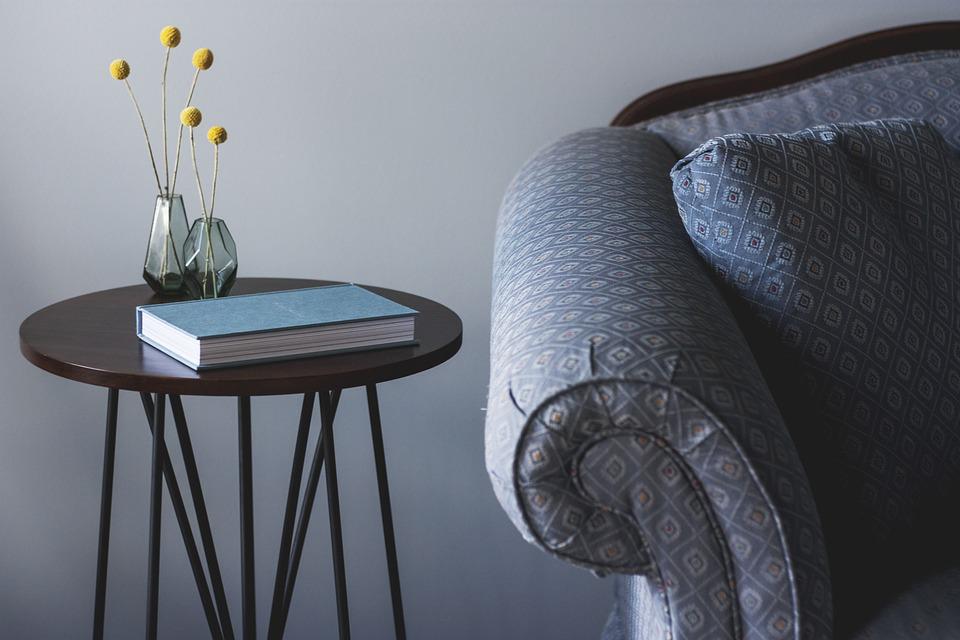 A imagem mostra um braço de sofá ao lado de uma mesinha com um vaso de flor e um livro, ilustrando um ambiente aconchegante