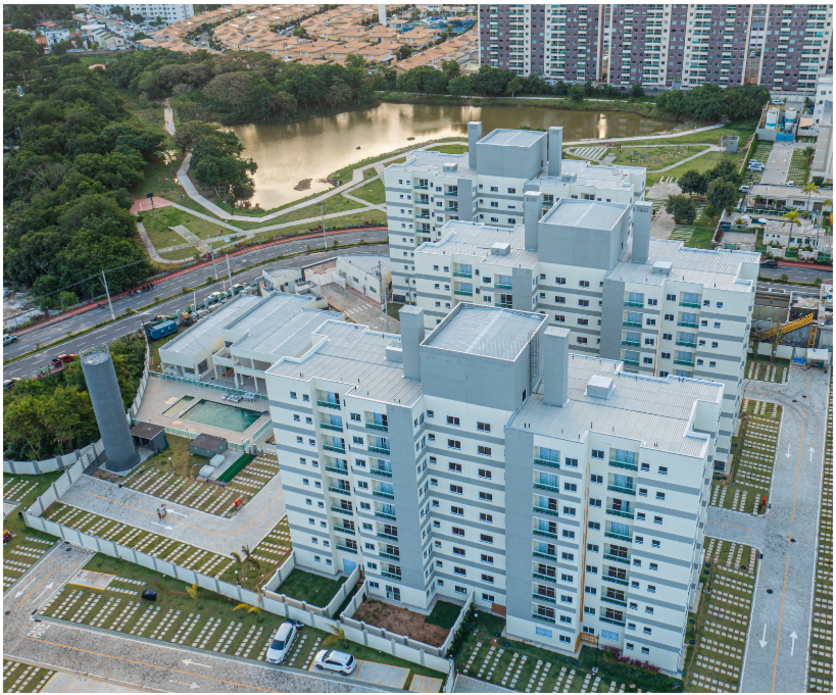 Vista aérea do empreendimento em Lauro de Freitas, Bahia. É possível ver o condomínio e também os arredores, como estacionamento, ruas, um lago e área verde.