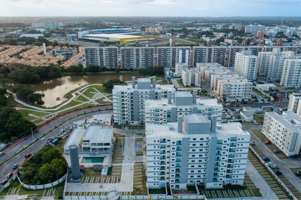 Vista aérea do empreendimento em Lauro de Freitas, Bahia. É possível ver o condomínio e também os arredores, como estacionamento, ruas, um lago e área verde.