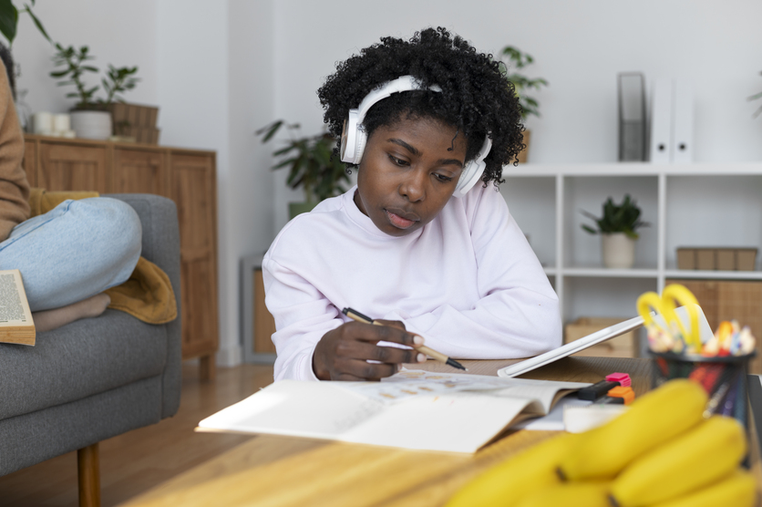 Jovem negra sentada à uma mesa de estudos, com um tablet, um livro e um caderno abertos.