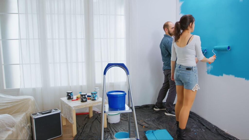 Um homem e uma mulher pintam uma parede interna da casa. Eles estão passando rolos de pintura na parede, a tinta é azul. Eles estão de costas e atrás deles é possível ver uma escada e uma banqueta com acessórios de pintura.