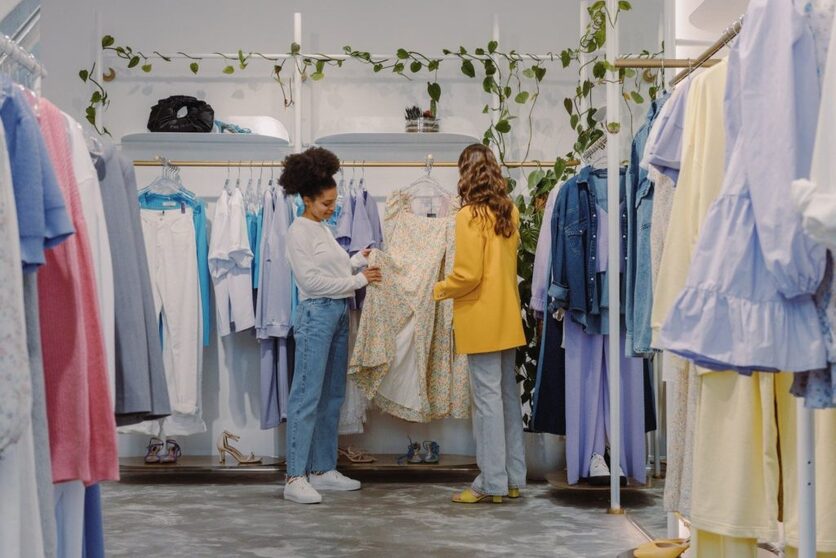 Duas mulheres numa loja de roupas escolhendo peças. À esquerda uma mulher negra com uma blusa branca, e à direita uma mulher branca com um casaco amarelo.