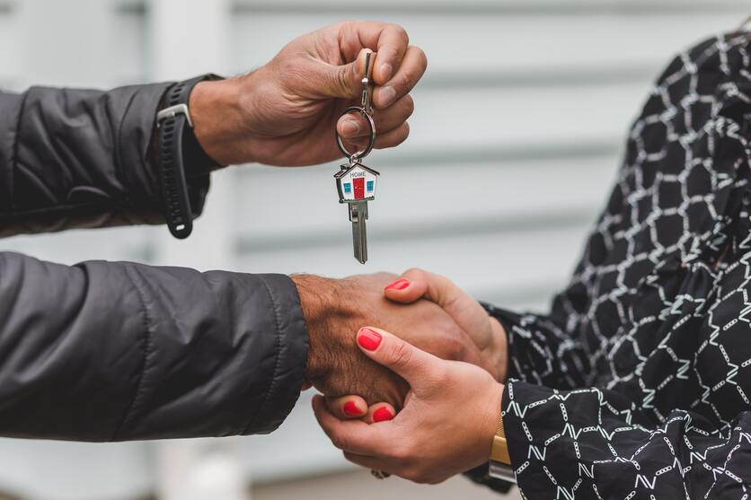 Um homem e uma mulher estão se cumprimentando, só é possível ver seus braços e as mãos entrelaçadas. Com a mão livre o homem segura uma chave com um chaveiro em formato de casa.