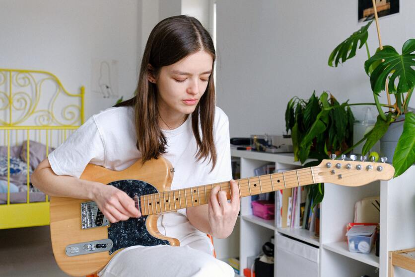 A imagem mostra uma moça segurando uma guitarra e sentada no meio do quarto para ilustrar o tempo livre para hobbies dentro de uma rotina organizada