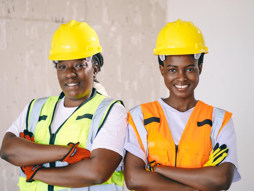 A imagem mostra duas mulheres com uniforme de construção com os braços cruzados e sorrindo para ilustrar a presença feminino na construção civil