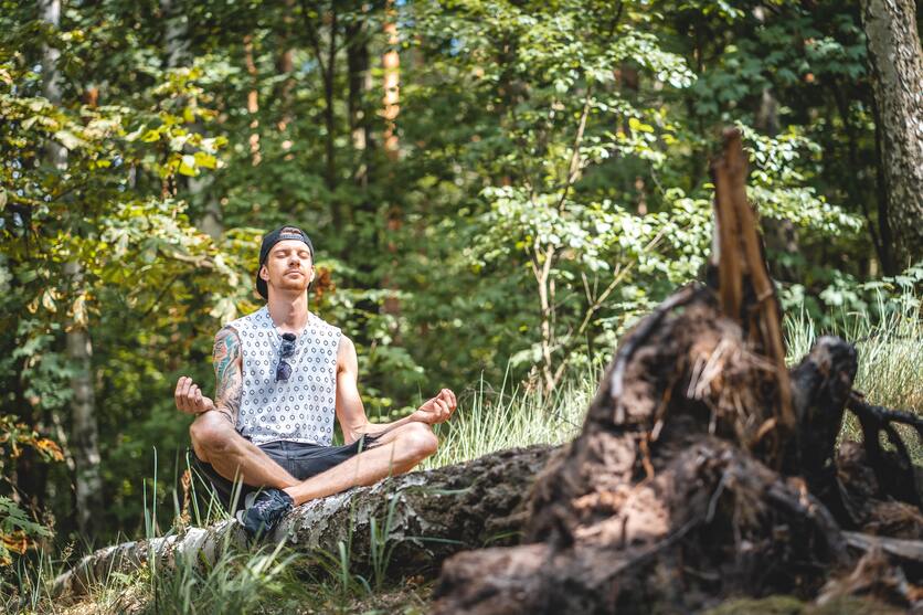 A imagem mostra um rapaz sentado em um tronco, no meio de uma área verde, em uma postura de meditação para ilustrar o que fazer na primavera