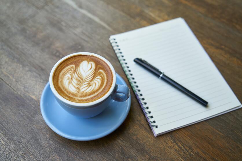 A imagem mostra uma xícara de café e um caderninho com uma caneta do lado para ilustrar a manhã de uma rotina organizada