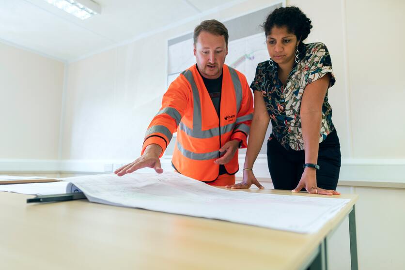 A imagem mostra um homem vestido com uniforme de construção e mostrando o projeto para uma mulher vestida de modo mais social para ilustrar a diversidade de profissionais para planejar uma empresa verde