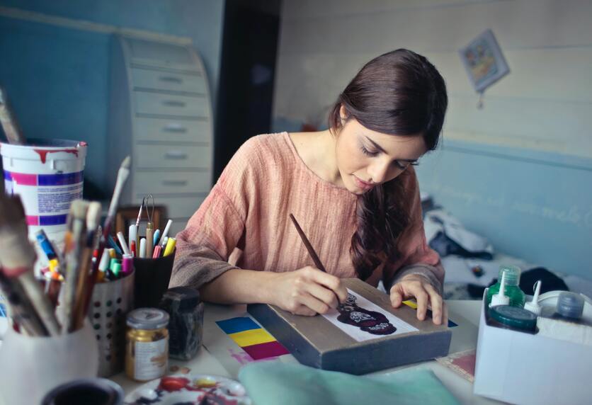 A imagem mostra uma mulher sentada e desenhando sobre um papel. Na mesa ao lado dela, está alguns potes com canetas e tintas, e ela segura um pincel para ilustar a importância de pausas no trabalho artístico