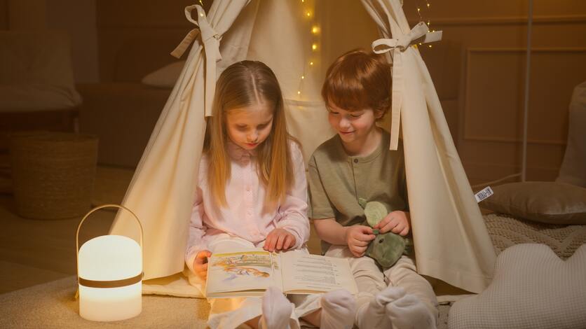 A imagem mostra duas crianças mais velhas, com cerca de 6 anos, lendo um livro embaixo de uma cabana para ilustrar a evolução do quarto montessoriano