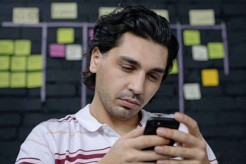A imagem mostra um homem olhado no celular, e ao fundo, está um painel cheio de post its para ilustrar a distração com o celular nas pausas do trabalho