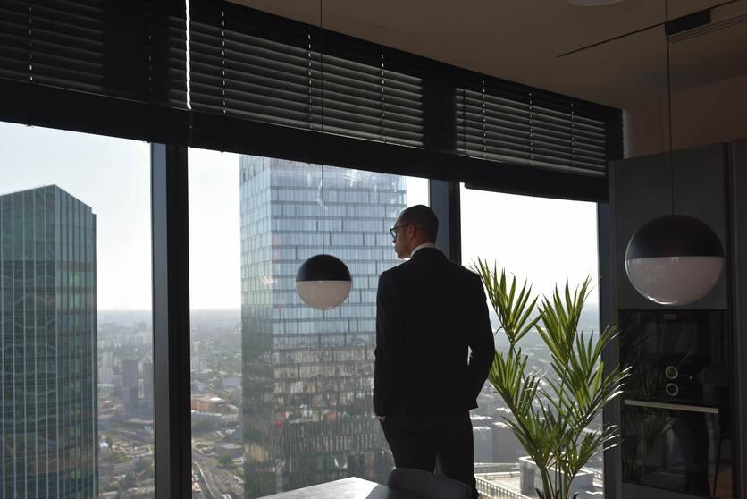 A imagem mostra um homem com terno em pé próximo a uma janela ampla e olhando a paisagem para ilustrar o que fazer nas pausas no trabalho