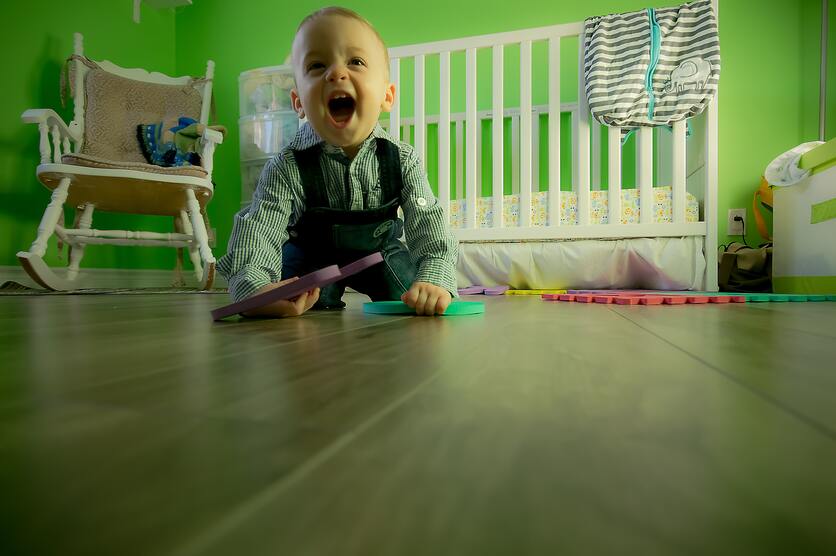 A imagem mostra um bebê engatinhando no chão e ao fundo é possível ver um berço com o colchão rente ao chão para ilustrar um berço montessoriano