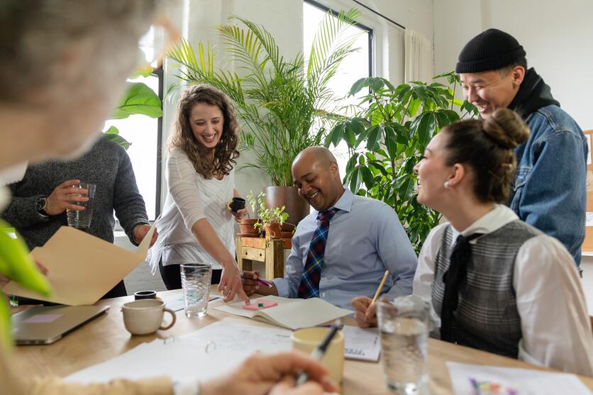 A imagem mostra uma mesa rodeada de pessoas com roupas de trabalho e sorrindo, na mesa estão copos de água, xícaras de café e folhas para ilustrar as pausas no trabalho