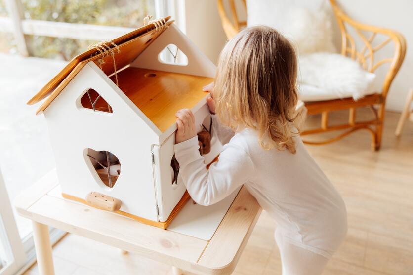 A imagem mostra uma criança se apoiando e m uma casinha de brinquedo que está em cima de uma mesinha para ilustrar os brinquedos no quarto montessoriano