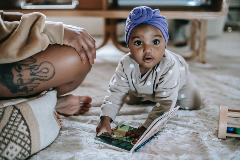 a imagem monstra um bebê sentado sobre os joelhos e com as mãos apoiadas em um livro no chão, com um grande tapete macio abaixo da criança para ilustrar as diversas texturas no quarto montessoriano