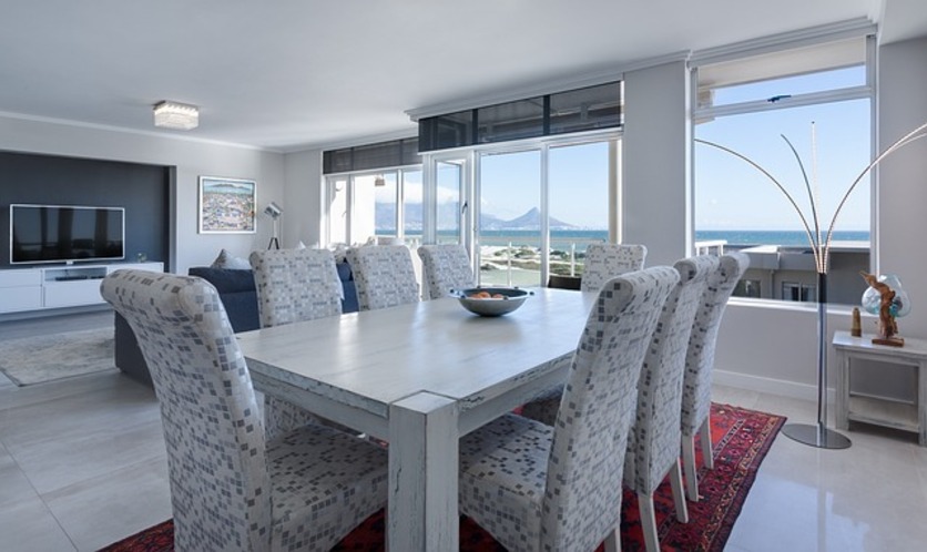 A imagem mostra uma sala de jantar com uma grande mesa de madeira, com cadeiras grandes e com tecido branco com ornamentos cinza. Há um tapete vermelho aparecendo embaixo da mesa e com grandes janelas ao fundo. Também é possível ver um ambiente de TV, com um sofá azul, um tapete cinza e uma TV ao fundo.