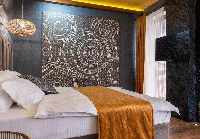 A imagem mostra um quarto com uma grande cama, com manta e vários travesseiros, além de paredes com texturas e pedras e um grande quadro para ilustrar o apartamento do signo de capricórnio