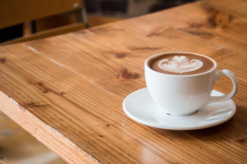 A imagem mostra uma mesa de madeira com uma xícara de café e pires, ambos brancos para contrastar com a madeira e com um desenho decorativo de espuma de leite sobre o café
