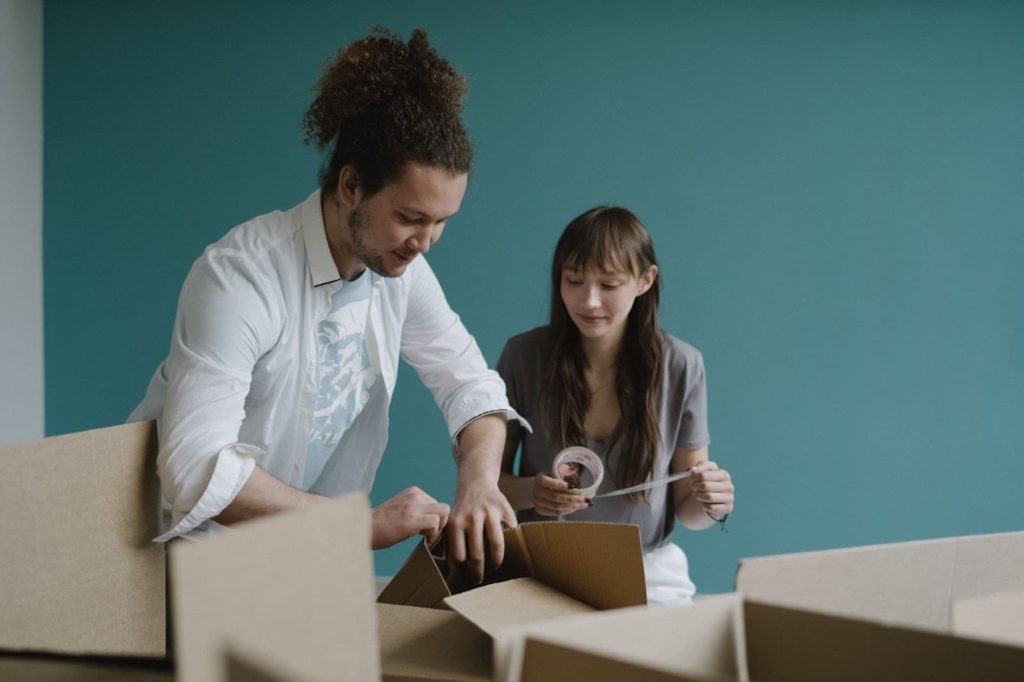 Imagem de duas pessoas, um homem e uma mulher, mexendo em caixas de mudança. A mulher segura uma fita durex para fechar as caixas