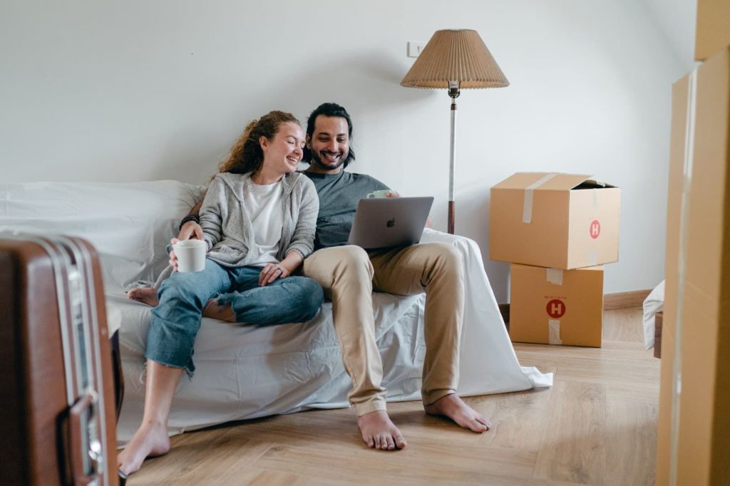 Imagem de um casal composto por um homem e uma mulher, sentados em um sofá ainda coberto e com caixas ao seu redor. Provavelmente são recém mudados para o apartamento