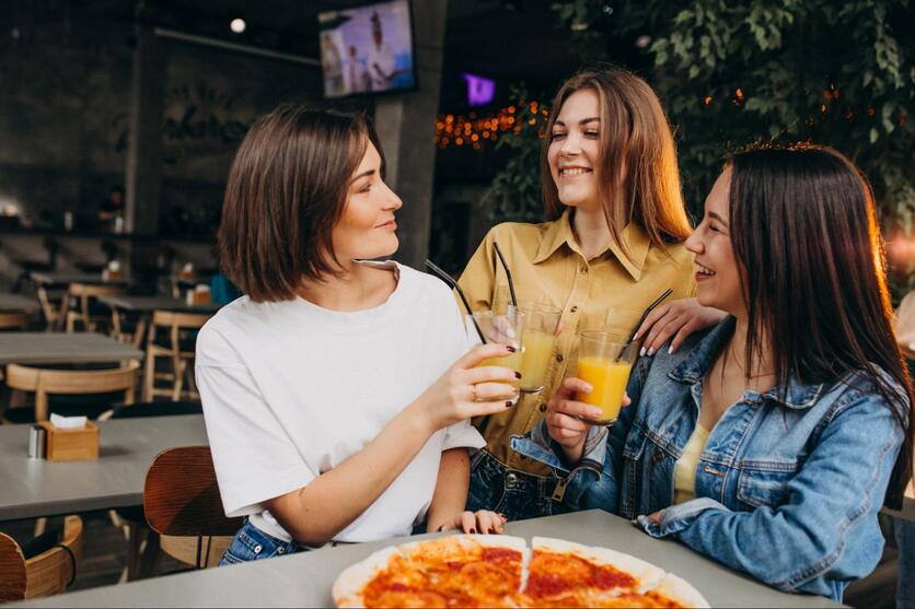 Três mulheres descontraídas, conversando e sorrindo, brindando em uma mesa de bar.