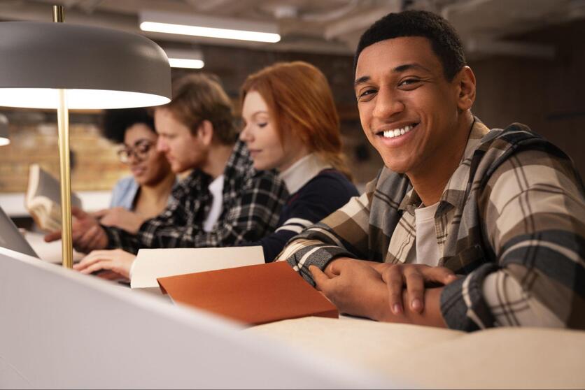 Jovem negro, sorrindo, sentado em uma mesa de estudos compartilhada.
