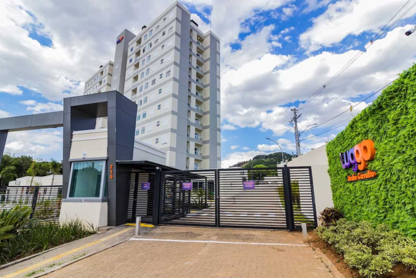 Fachada do condomínio Luggo Jardim Carvalho, destacando o portão de entrada e um edifício de vários andares ao fundo sob um céu parcialmente nublado.