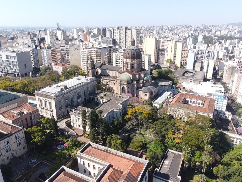 Vista panorâmica do centro histórico de Porto Alegre, mostrando edifícios históricos, incluindo uma catedral com cúpula, rodeada por áreas verdes e a paisagem urbana ao fundo.