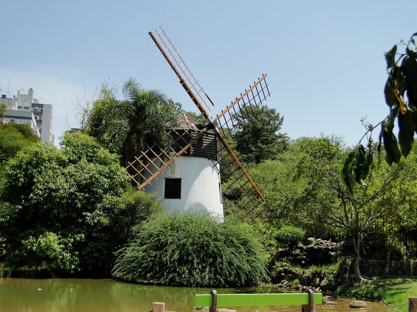 Vista do moinho de vento no Parque Parcão, no bairro Moinhos de Vento, cercado por vegetação densa e um lago tranquilo, sob um céu claro.