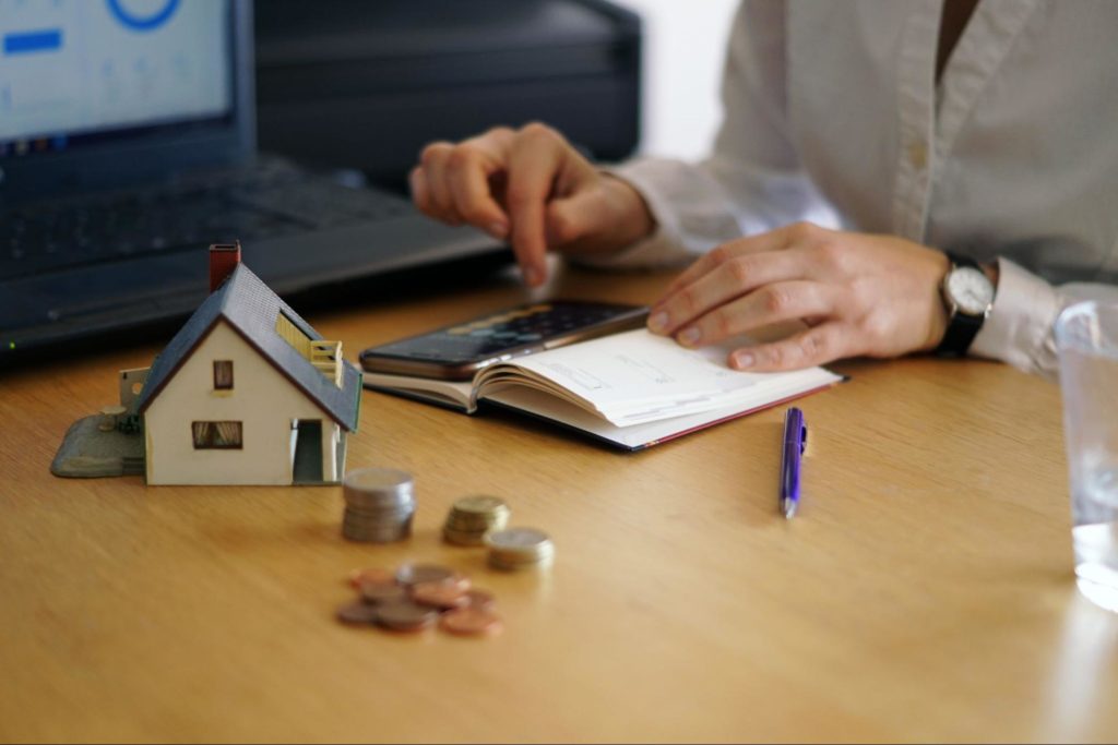 Proprietário pensando em soluções para inadimplência, fazendo anotações e contas numa calculadora, com uma miniatura de casa e moedas na mesa.