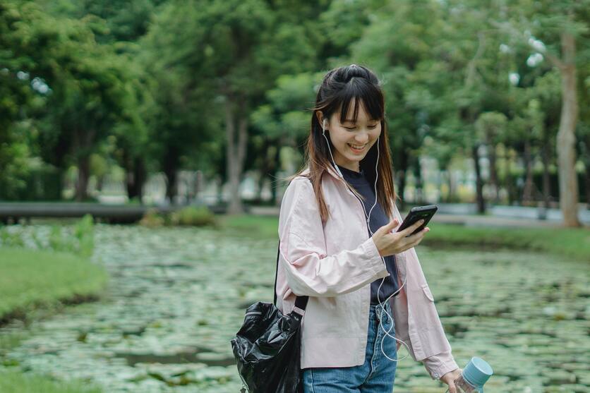 Mulher andando por um parque durante o dia, sorrindo, olhando o celular.