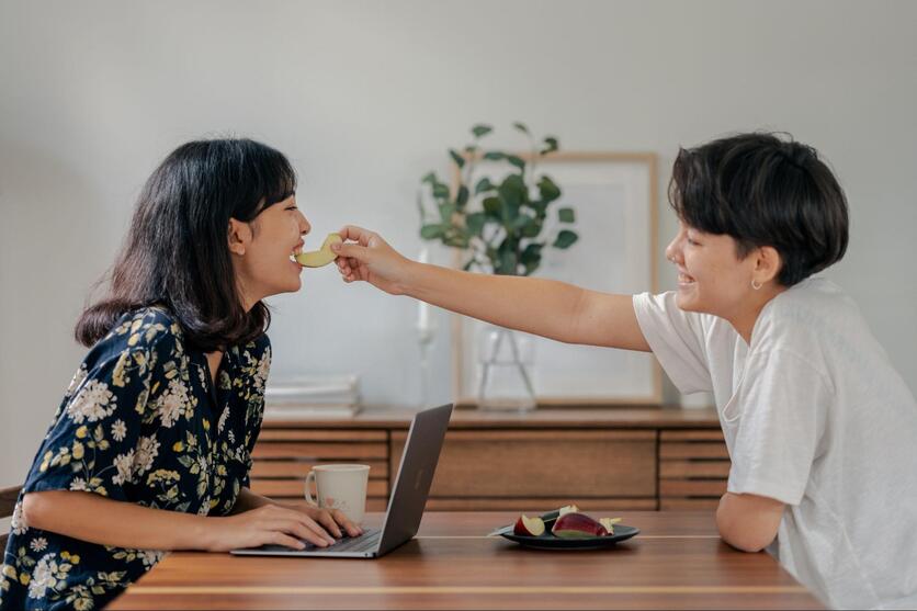 Mulher sorrindo em sua mesa de trabalho home office enquanto parceira a alimenta com frutas. 