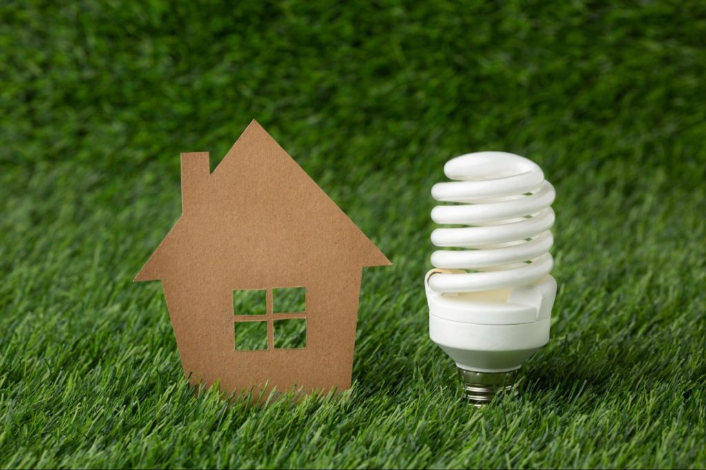 Papel recortado em formato de uma casa ao lado de uma lâmpada, ambos na grama, para representar sustentabilidade.