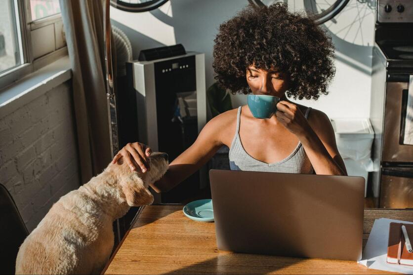 Mulher negra na frente do computador, tomando café e fazendo carinho em seu cachorro.