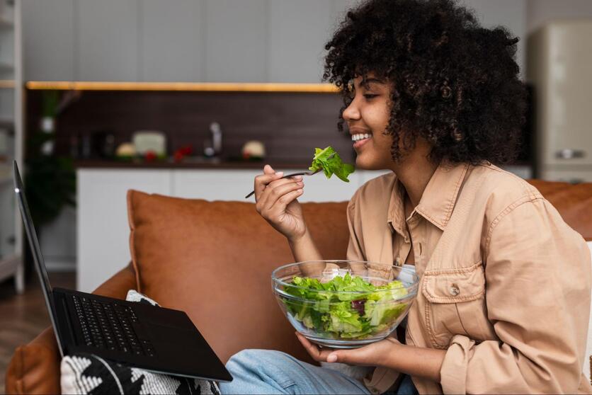 Mulher negra sorrindo, sentada no sofá enquanto come uma salada de microverdes.