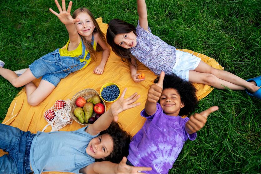 Quatro crianças deitadas na grama, sorrindo. No meio, há uma toalha de piquenique com cestas de frutas.