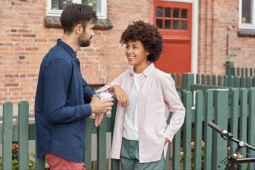 Mulher negra, sorrindo enquanto conversa com homem em frente à uma casa.