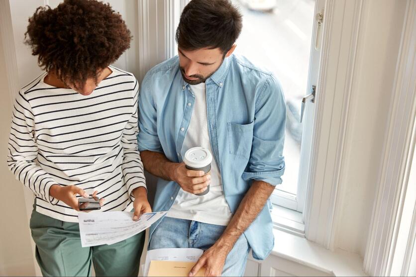 Casal sentado à janela de uma casa, analisando papéis juntos.