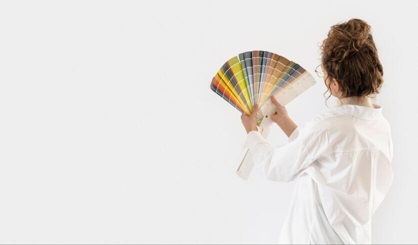 Mulher de costas com diversas amostras de cores em mãos, analisando as cores e a parede.