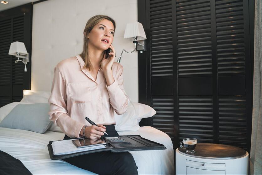 Mulher com roupa social, sentada na cama de um flat, com uma pasta de trabalho no colo enquanto atende uma chamada no celular.