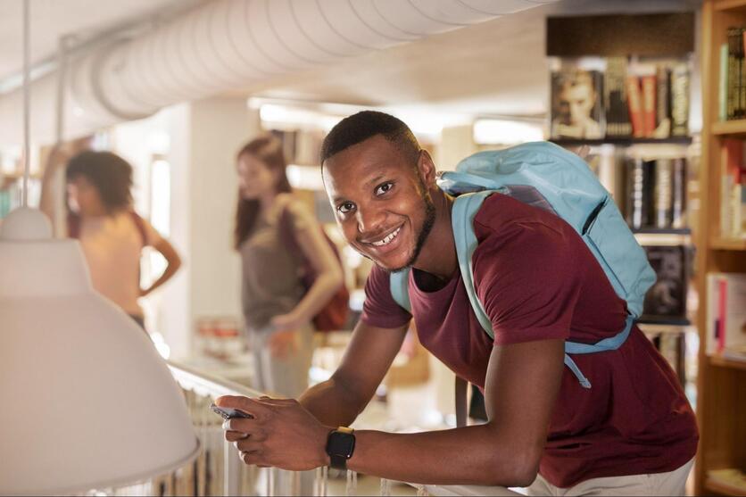 Jovem estudante negro, sorrindo, mexendo no celular dentro de uma biblioteca universitária.