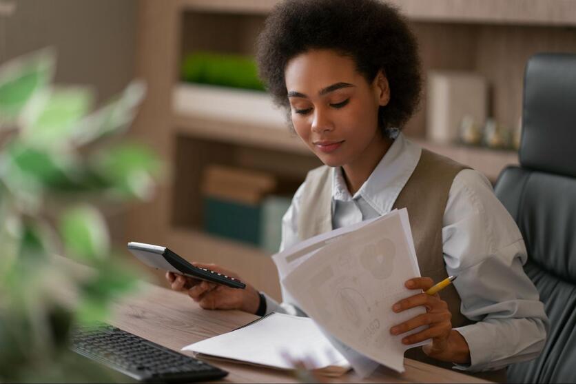 Mulher negra, sentada em frente ao computador, segurando documentos em uma mão e uma calculadora na outra.