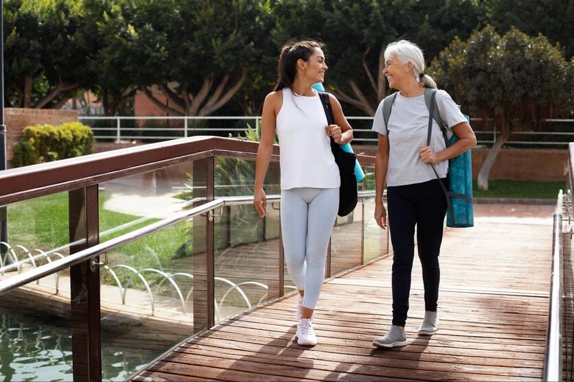 Duas mulheres adultas, caminhando por um parque, sorrindo e conversando enquanto carregam bolsas com material de ioga.