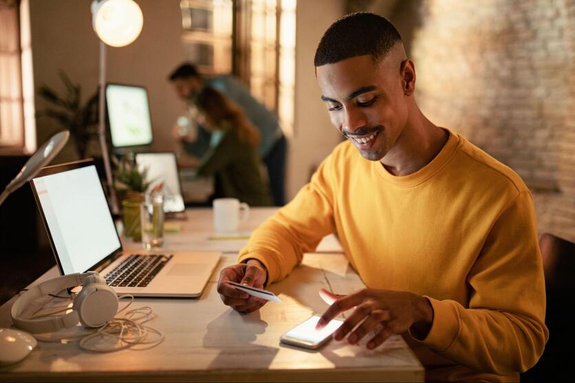 Jovem negro sentado em frente ao computador, sorrindo, com um cartão de crédito em mãos enquanto mexe no celular.